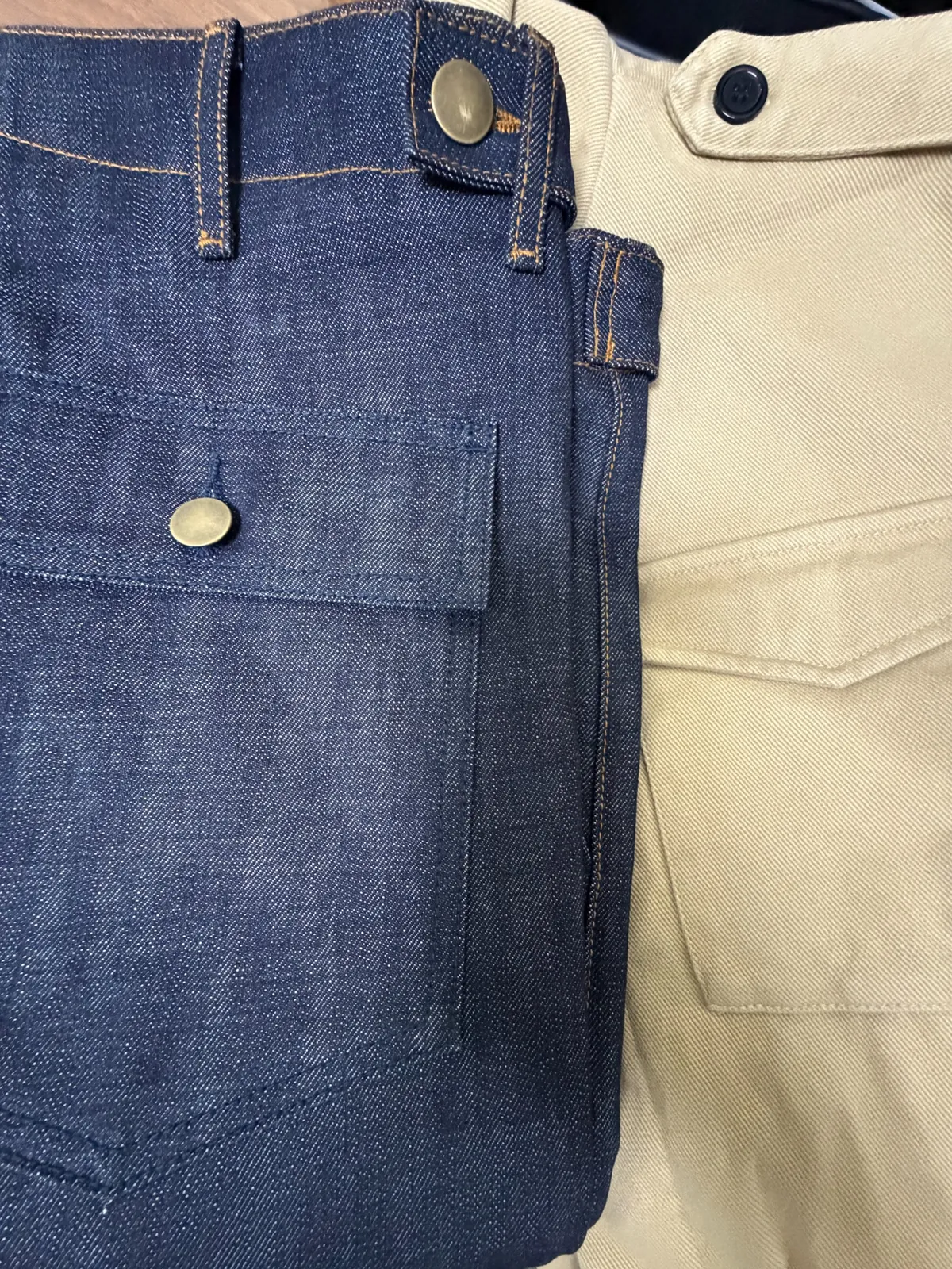 Close-up of indigo denim pocket, contrast stitching and brass-tone metal buttons beside a khaki garment